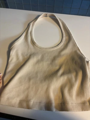 Ribbad beige linnetopp - Säljer en ribbad beige linnetopp med halterneck och croppad passform. Toppen har smala ränder och är ärmlös, perfekt för varma dagar eller som layering-piece. Enkel och stilren design som passar till mycket.