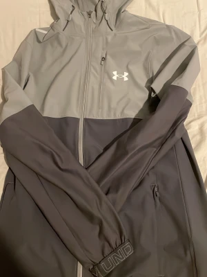 Grå vindjacka från Under Armour - Snygg tvåfärgad vindjacka från Under Armour i ljusgrått och mörkgrått. Jackan har huva, hel dragkedja, bröstficka med dragkedja och två sidofickor. Tillverkad i ett lätt och vindtätt material, perfekt för blåsiga dagar.