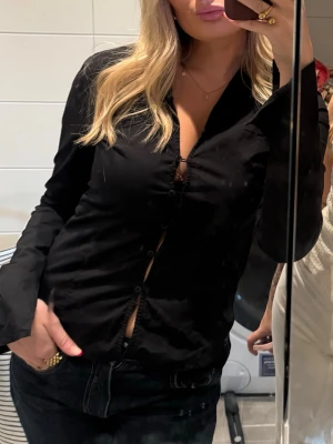 Svart skjorta - Säljer denna stilrena svarta skjorta som är ett riktigt statement-plagg! Den har en figurnära passform som framhäver silhuetten på ett snyggt sätt och funkar lika bra uppknäppt över ett linne som knäppt för en mer dressad look.