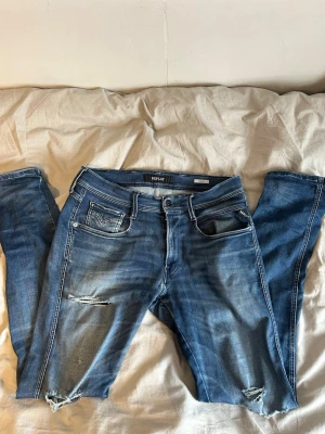 Blå slitna jeans från Replay - Tvär feta blå jeans från Replay med slitningar och hål på benen för en trendig look. Klassisk femficksmodell med dragkedjegylf och sköna stretchiga material. Perfekta för dig som gillar en avslappnad och modern stil.