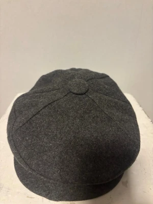 Klassisk Flat Cap från Norcap – Norsk design & Paisleyfoder - Klassisk gubbkeps från norska Norcap – Storlek 56 Säljer en mycket fin och välvårdad keps (flat cap) från det norska märket Norcap (Opplandske Luefabrikk A/S). Kepsen är tillverkad i Gjøvik, Norge, och håller en mycket hög kvalitet.  Material: Kraftig och värmande ullmix i en snygg mörkgrå melerad färg.  Design: Klassisk modell med tryckknapp på skärmen.  Insida: Otroligt snyggt och unikt paisleymönstrat foder i rött, blått och gult som ger en lyxig känsla.  Storlek: 56.  Skick: Gott skick.
