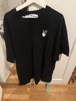 Svart Off-White t-shirt - Säljer en svart t-shirt från Off-White med liten vit logga och handdetalj på bröstet. Klassisk rund halsringning och korta ärmar. Tillverkad i mjuk bomull för en skön och avslappnad look.