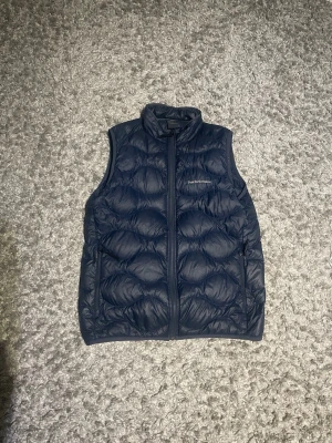 Mörkblå dunväst Peak Performance - Snygg mörkblå dunväst från Peak Performance med hög krage och dragkedja framtill. Västen har ett quiltat mönster och två sidofickor. Perfekt att slänga över en hoodie eller tröja för extra värme. Materialet är lätt och smidigt, vilket gör den enkel att bära. Storlek 170 men passar S, jag själv har S-M. Pris går diskutera 