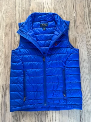 Blå dunväst från Polo Ralph Lauren - Snygg blå dunväst från Polo Ralph Lauren med quiltad design och två dragkedjeförsedda fickor framtill. Västen har hög krage och den klassiska röda loggan broderad på bröstet. Perfekt för lager-på-lager och riktigt stilren look.