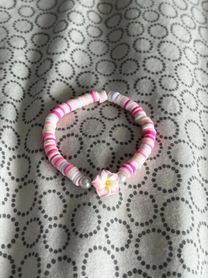 Pärlarmband med rosa blomma - Sött armband med platta pärlor i olika rosa och vita nyanser samt två silverfärgade pärlor. I mitten sitter en dekorativ blomma i rosa, vitt och gult. Armbandet är elastiskt och har en lekfull, färgglad design.