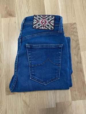 Jacob choën jeans - Säljer ett par mörkblå skinny jeans med en unik, broderad patch i svart, vitt och rött på linningen. Jeansen har klassisk femficksdesign och är tillverkade i stretchigt denimtyg för en bekväm passform. Perfekta till din vardagsstil! 