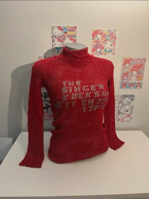 Röd glittrig stickad polotröja - Säljer en röd stickad polotröja med glittereffekt och ett silvrigt texttryck framtill. Tröjan har lång ärm, ribbad struktur och figurnära passform. Perfekt för dig som vill sticka ut med en trendig och bekväm look.
