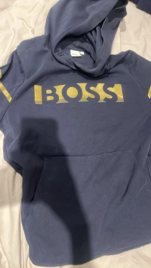 Mörkblå hoodie från BOSS med guldtryck. Priset kan diskuteras.  - Snygg mörkblå hoodie från BOSS med stort guldigt BOSS-tryck på bröstet och gulddetaljer på ärmarna. Klassisk känguruficka framtill och stor huva. Perfekt för dig som vill ha en stilren men ändå iögonfallande look. Mjuk och skön bomullskänsla.