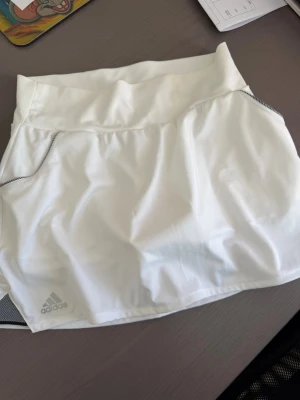 Vit sportkjol från Adidas Climalite - Snygg vit kortkjol från Adidas med Climalite-material som andas. Kjolen har diskreta grå detaljer vid sidorna och en bred midjeresår för skön passform. Perfekt för tennis eller andra sportaktiviteter. Adidas-logga nere till vänster. Aldrig använd 