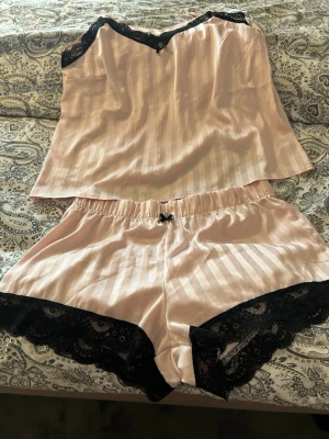 Rosa satinpyjamas med spetsdetaljer - Säljer ett tvådelat pyjamasset i ljusrosa satin med svarta spetsdetaljer. Linnet har tunna axelband och en liten rosett framtill, medan shortsen har elastisk midja, svarta spetskanter och en söt rosett. Perfekt för mysiga kvällar hemma. Storlek Xl men jätte liten i storleken så skulle säga Medium