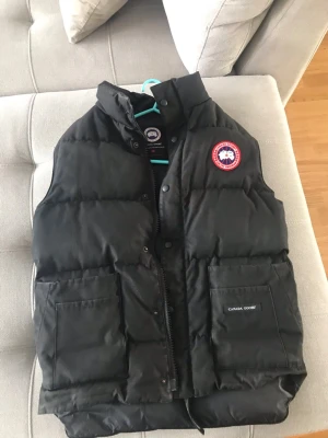 Svart dunväst från Canada Goose - Säljer en svart dunväst från Canada Goose med klassisk logga på bröstet. Västen har hög krage, två stora fickor framtill och dragkedja med knappar. Perfekt för lager-på-lager och riktigt varm tack vare dunfyllningen. Ganska stor för att vara xs 