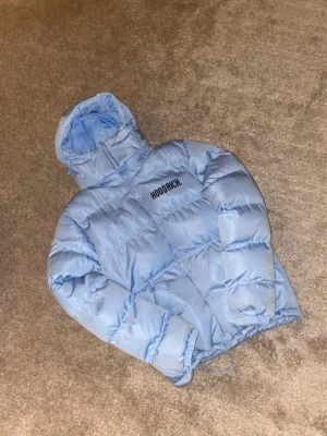 Ljusblå pufferjacka från Hoodrich - Snygg ljusblå pufferjacka från Hoodrich med huva och dragkedja framtill. Jackan har ett puffigt, quiltat material och Hoodrich-loggan tryckt på bröstet. Perfekt för kalla dagar när du vill hålla stilen och samtidigt vara varm.