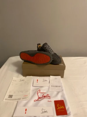 Christian Louboutin grå sneakers - ÄKTA Snygga grå sneakers från Christian Louboutin i mocka med ikoniska röda sulor och coola nitar på tån. Skorna har rund tå, platt sula och grå snörning. Perfekta för dig som vill sticka ut med exklusiv streetstyle. Tre  nitar saknas, Kvitto finns
