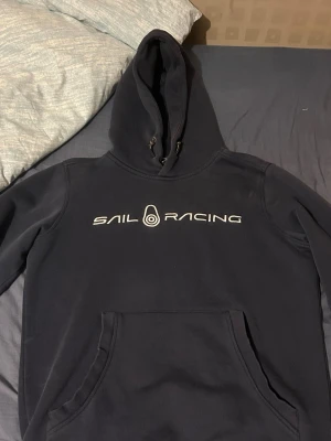 Marinblå Sail Racing hoodie - Snygg marinblå hoodie från Sail Racing med stor vit logga tryckt över bröstet. Klassisk känguruficka framtill och justerbar huva med snören. Perfekt för dig som gillar sportig och avslappnad stil. Mjuk och skön bomullsblandning.