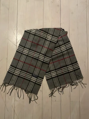 Grå rutig halsduk från Burberrys - Priset går att diskutera Klassisk grå halsduk från Burberrys i 100% lammull. Halsduken har ett rutigt mönster i svart, vitt och rött samt fransar i kanterna. Perfekt accessoar för att lyfta din outfit med en tidlös brittisk stil. Rikigt bra till dåligt väder 