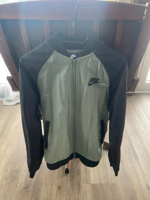 Nike bomberjacka grå och svart - Nike bomberjacka i grått med svarta ärmar och detaljer. Klassisk bombermodell med dragkedja framtill, ribbade muddar och två fickor. Nike-logga broderad på bröstet. Tillverkad i ett lätt, glansigt material som ger en sportig vibe.
