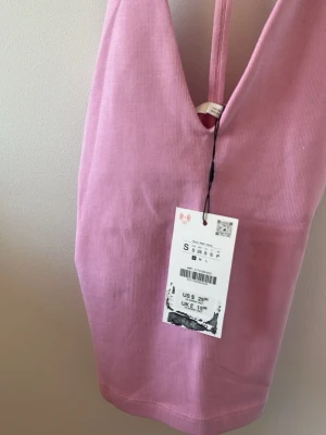 Ljusrosa ribbad linnetopp - Säljer en ljusrosa ribbad linnetopp med smala axelband och v-ringad hals. Toppen har en croppad passform och är perfekt för varma dagar. Enkel och stilren design som är lätt att matcha.