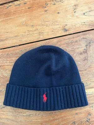 Mörkblå mössa från Polo Ralph Lauren - Snygg mörkblå ribbstickad mössa från Polo Ralph Lauren med klassisk röd broderad logga framtill. Mössan har uppvikt kant och är tillverkad i mjukt stickat material som håller värmen. Perfekt accessoar för kalla dagar.
