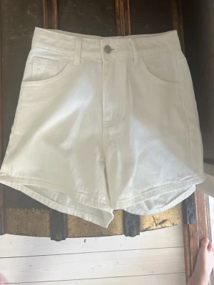 Vita högmidjade shorts i bomull - Snygga vita shorts med hög midja och klassisk femficksdesign. Tillverkade i mjuk bomull med raka ben och bälteshällor. Perfekta för varma dagar och enkel att matcha med olika toppar.