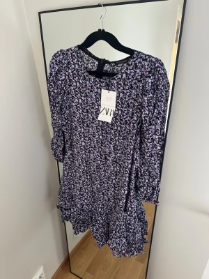 Blommig kortklänning från Zara - Supersöt kortklänning från Zara med svart botten och små lila blommor. Klänningen har långa ärmar, volangdetaljer nertill och en rund halsringning. Perfekt för dig som gillar romantiska och trendiga plagg.