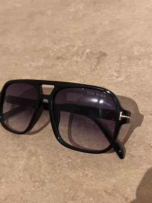Svarta Tom Ford pilotbågar solglasögon - Snygga svarta solglasögon från Tom Ford med klassisk pilotbåge och mörka tonade glas. Bågarna har en cool detalj med guldfärgad T-logga vid gångjärnen. Perfekta för dig som vill ha en stilren och trendig look.