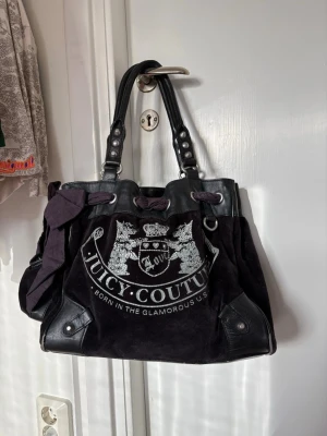 Svart Juicy Couture handväska - Cool svart handväska från Juicy Couture med stort silverfärgat logotryck framtill. Väskan har dubbla handtag i läder, dekorativ rosett och detaljer i mocka och läder. Perfekt för dig som gillar statement-accessoarer och klassisk Y2K-stil.
