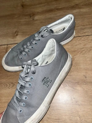 Gråa Tommy Hilfiger sneakers - Snygga grå sneakers från Tommy Hilfiger med klassisk låg modell. Skorna har canvasmaterial, vita sulor och gråa skosnören. På sidan finns TH-loggan broderad i silver. Perfekta för en clean och stilren look.