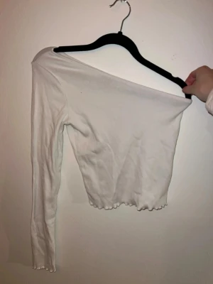 Vit oneshoulder topp Gina Tricot - Trendig vit oneshoulder topp från Gina Tricot i ribbad bomullsmix. Croppad modell med lång ärm och vågig kant nertill för en extra snygg detalj. Det är storlek xs