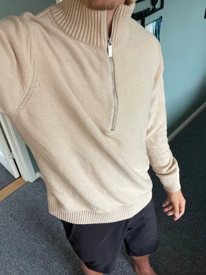 Beige stickad halvzip tröja selected home - Säljer en beige stickad tröja med halvzip och hög ribbad krage. Tröjan har långa ärmar och ribbade muddar vid ärmslut och nederkant. Perfekt för lager på lager och enkel att matcha med olika outfits.
