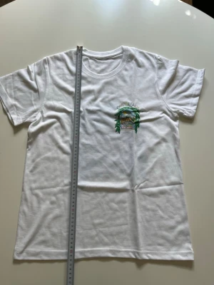 Vit Casablanca t-shirt med tryck - Vit t-shirt i bomull med rund hals och korta ärmar. På bröstet finns ett färgglatt tryck med naturmotiv och texten 'Green Place'. Perfekt för en avslappnad och stilren look. Storlek M passar S, nyskick.