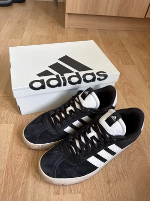 Svarta Adidas sneakers med vita ränder - Stilrena svarta Adidas sneakers med klassiska vita ränder på sidorna och vit plös. Ovandelen är i mocka och skon har svarta skosnören. Skorna är sparsamt använda och det syns på sulan och mockan. Skorna är i ett fint skick och fungerar exakt som de ska, utan defekter eller större skador. Skriv gärna vid funderingar eller frågor!