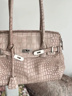 Beige handväska i krokodil - Köpt second hand så den har den valiga secondhand lukten! Säljer en beige handväska i krokopräglat läder med silverfärgade metalldetaljer och dubbla handtag. Väskan har klassisk rektangulär form och dekorativa spännen framtill. Perfekt för dig som gillar en lyxig och trendig look.