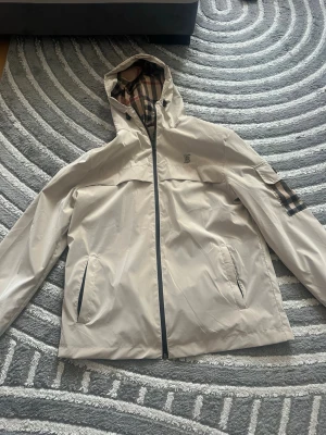 Beige vindjacka från Burberry - Snygg beige vindjacka från Burberry med klassiskt rutigt foder i huvan och på ärmen. Jackan har dragkedja framtill, två fickor med dragkedja och en patch med Burberry-logga på ärmen. Tillverkad i ett lätt och vattenavvisande material.