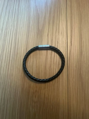 Svart flätat läderarmband - Stilrent svart armband i flätat läder med ett enkelt magnetlås i silverfärgad metall. Perfekt accessoar för en clean och modern look. Passar både till jeans och skjorta för en avslappnad men ändå snygg stil.