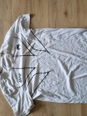 Vit UFC t-shirt med svart tryck - Säljer en vit t-shirt från UFC med svart grafiskt tryck och logga på bröstet. T-shirten har rund hals och korta ärmar, perfekt för dig som gillar sportig och clean stil.