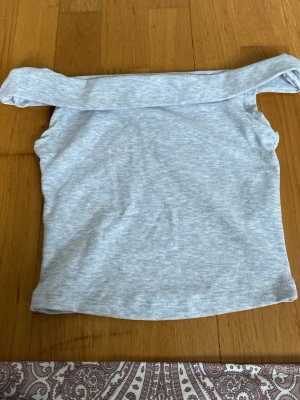 Ljusgrå offshoulder topp Bershka - Trendig ljusgrå offshoulder topp från Bershka. Toppen är ärmlös och har en bred kant upptill som ger en snygg och modern look. Perfekt att matcha med jeans eller kjol för en cool stil.