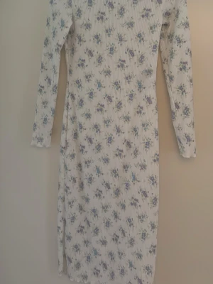 Vit blommig långärmad klänning nyskick!  - Söt vit långärmad klänning med små blå och lila blommor. Klänningen har en ribbad struktur och är figurnära med en slits på sidan. Perfekt för vår och sommar med sitt romantiska blommönster. Nyskick! 