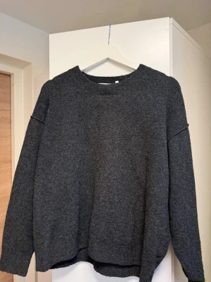 Mörkgrå oversized stickad tröja - Mysig mörkgrå stickad tröja med avslappnad, oversized passform och rund halsringning. Tröjan har långa ärmar och ribbade muddar vid hals, ärmslut och nederkant. Perfekt för lager på lager och chill dagar.
