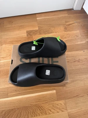 Svarta Yeezy Slide tofflor - Säljer ett par svarta Yeezy Slide tofflor från Adidas. De har en chunky, modern design med öppen tå och räfflad sula för extra grepp. Tillverkade i mjukt, flexibelt syntetmaterial som ger skön komfort. Perfekta för chill dagar hemma eller ute.