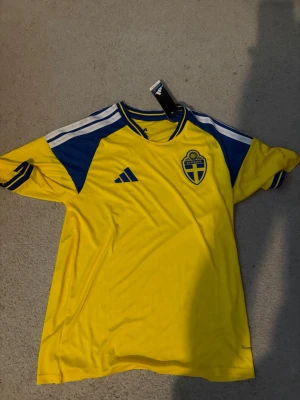 Svensk landslagströja Adidas gul/blå - Säljer en gul och blå svensk landslagströja från Adidas med klassiska tre ränder på axlarna och landslagets emblem på bröstet. Tröjan har rund hals och är tillverkad i ett lätt, ventilerande material. Perfekt för match eller träning!