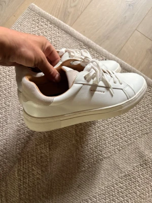 Vita sneakers från Björn Borg - Stilrena vita sneakers från Björn Borg i läder med klassisk låg profil och vita snören. Skorna har diskret logga på sidan och bak på hälen, samt beige detaljer vid ankeln. Perfekta för en clean och modern look.