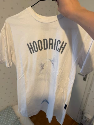 Vit HOODRICH t-shirt med tryck - Säljer en vit t-shirt från HOODRICH med svart texttryck framtill och motiv av duvor både fram och bak. På ryggen finns även blå text och ett större duvmotiv. T-shirten har rund hals och korta ärmar, perfekt för en avslappnad streetwear-look.