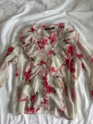 Blommig volangblus från ZARA - Supersöt blus från ZARA i vitt med rosa blommönster och volangdetaljer längs fram- och halsringning. Blusen har långa ärmar och är gjord i ett tunt, transparent material som känns luftigt och romantiskt. Perfekt för dig som gillar en feminin och lekfull stil. Blusen är i storlek S och postas och packas varsamt!💓