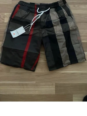 Randiga shorts med snörning - Snygga shorts med breda ränder i svart, beige, rött och mörkgrått. De har elastisk midja med vit snörning och avslappnad passform. Perfekta för sommaren och har en modern streetwear-vibe. Materialet känns lätt och bekvämt.