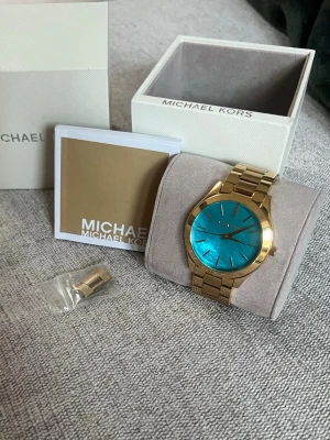 Guldig Michael Kors klocka turkos urtavla - Snygg klocka från Michael Kors med guldig länk och en iögonfallande turkos urtavla. Klockan har klassisk rund form och levereras med originalask, manual och extra länkbit. Perfekt accessoar för dig som vill sticka ut. Ge prisförslag 