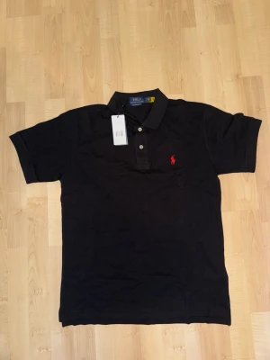 Svart pikétröja från Polo Ralph Lauren - Klassisk svart pikétröja från Polo Ralph Lauren med korta ärmar och krage. Tröjan har två knappar framtill och den ikoniska röda logotypen broderad på bröstet. Tillverkad i bomull för en bekväm och stilren look.