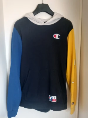 Champion hoodie limited edition - Snygg hoodie från Champion med svart kropp, blått ärm, gul ärm och vit huva. Klassisk Champion-logga på bröstet och lappen nertill. Mjuk bomullsmix och känguruficka framtill. Perfekt för dig som gillar streetwear och färg. Funkar för varjedag och sport. 