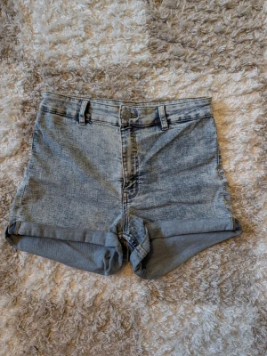 Högmidjade ljusblå jeansshorts - Säljer ett par högmidjade ljusblå jeansshorts med uppvikta benslut och bälteshällor. Klassisk femficksmodell och stängs med knapp och dragkedja. Perfekta till sommaren och lätta att matcha med olika toppar.