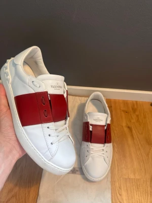 Valentino vita sneakers med röd detalj - Snygga vita sneakers från Valentino med en bred röd läderdetalj över vristen. Klassisk låg modell med vita snören och diskret logga på plösen. Skorna har en stilren och modern design i läder som passar perfekt till streetwear.