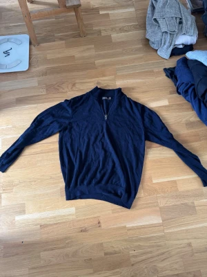 Mörkblå halvzip från Oscar Jacobson - Mörkblå stickad tröja från Oscar Jacobson med halv dragkedja och ribbade muddar. Klassisk modell med hög krage, perfekt för lager på lager. Materialet känns mjukt och följsamt, vilket gör den skön att bära till jeans eller chinos. Små hål som visat men annars bra. 